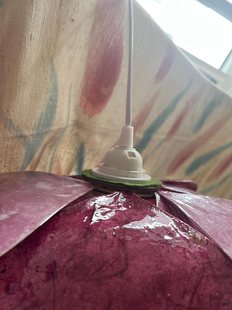 May include: A pendant lamp shade featuring overlapping dark magenta, textured, fibrous petals arranged in a circular pattern. The shade is attached to a white plastic socket and a white electrical cord, with a green felt ring spacer.