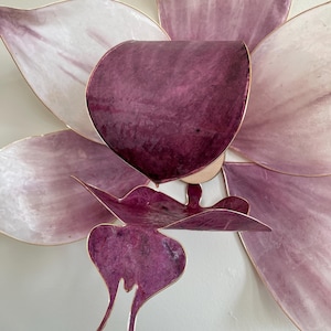 May include: A wall-mounted flower sculpture featuring large, translucent petals in shades of pale pink and deep purple. The structure is defined by thin, metallic gold-toned edges that outline each petal, creating a layered, dimensional floral form.