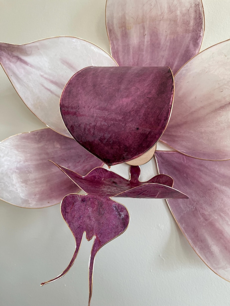 May include: A wall-mounted flower sculpture featuring large, translucent petals in shades of pale pink and deep purple. The structure is defined by thin, metallic gold-toned edges that outline each petal, creating a layered, dimensional floral form.