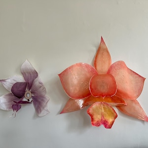 May include: Two wall-mounted flower sculptures. The larger flower features translucent, layered petals in shades of orange, coral, and yellow. The smaller flower displays translucent, layered petals in soft shades of purple and white with a central ring.
