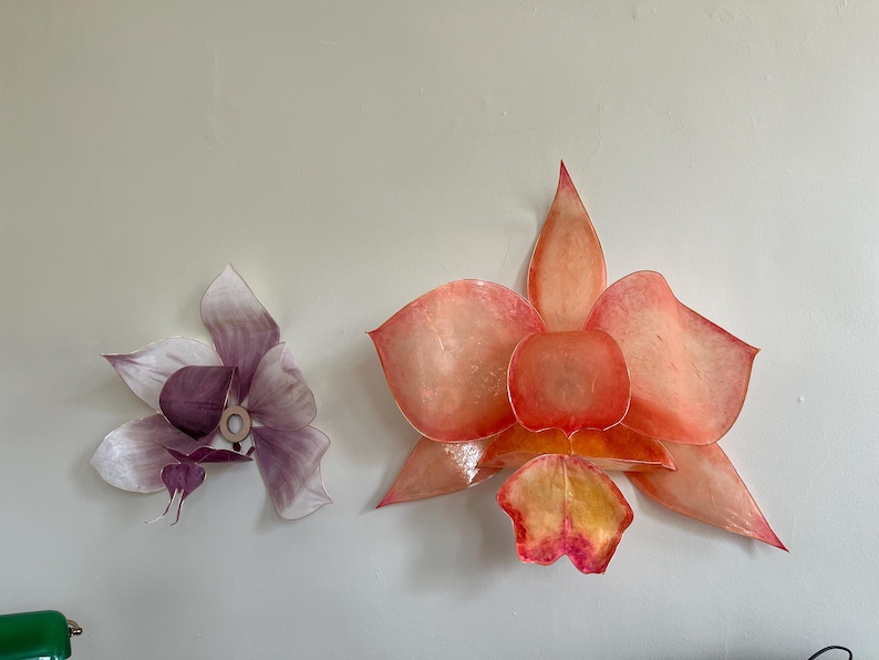 May include: Two wall-mounted flower sculptures. The larger flower features translucent, layered petals in shades of orange, coral, and yellow. The smaller flower displays translucent, layered petals in soft shades of purple and white with a central ring.