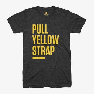 May include: Dark gray t-shirt with the words "PULL YELLOW STRAP" printed in yellow. The text is arranged vertically, with a horizontal yellow line beneath the word "STRAP". The shirt has a crew neck and short sleeves.