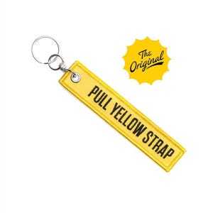 Puede incluir: Llavero amarillo con el texto "PULL YELLOW STRAP" en negro. La etiqueta tiene un anillo de metal y un pequeño sol amarillo con las palabras "The Original" en negro.