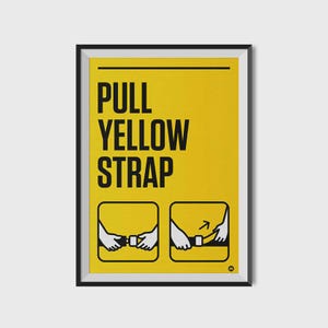 May include: A framed poster with a bright yellow background and black text that reads "PULL YELLOW STRAP." Below the text are two illustrated diagrams showing how to use a seatbelt. The poster is in a black frame.