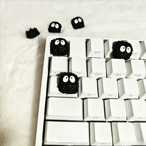 Op de afbeelding: Een wit mechanisch toetsenbord met verschillende zwarte, fuzzy keycaps in de vorm van cartoon roet sprites. De keycaps hebben grote, witte ogen. Het toetsenbord ligt op een witte, zachte ondergrond.