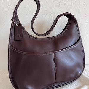 Op de afbeelding: Een donkerbruine leren schoudertas met een gebogen vorm en een enkele schouderriem. De tas heeft een voorvak en een klein leren label aan de riem. De tas is ongeveer 30 cm breed en 25 cm hoog.