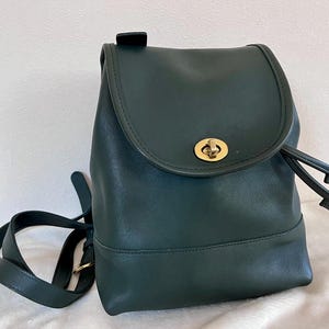 May include: A dark green leather backpack with a flap closure and a gold-colored turn lock. The bag has a drawstring closure and a single adjustable shoulder strap. The backpack is approximately 10 inches tall.