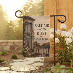 May include: A rectangular garden flag hanging on a black metal stand, featuring a coastal scene with a weathered door, a white picket fence, and the text SALT AIR and the RUST on YOUR DOOR printed in black serif font against a soft beige background.
