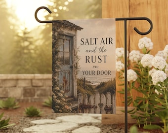 May include: A rectangular garden flag hanging on a black metal stand, featuring a coastal scene with a weathered door, a white picket fence, and the text SALT AIR and the RUST on YOUR DOOR printed in black serif font against a soft beige background.
