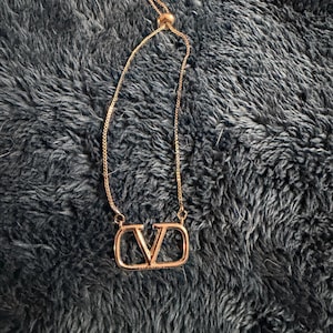 Puede incluir: Un collar de tono oro rosa que presenta un colgante central con forma de letra V dentro de un marco rectangular. La pieza incluye una cadena delicada con un mecanismo de cuenta deslizante ajustable, lo que permite un ajuste personalizado.