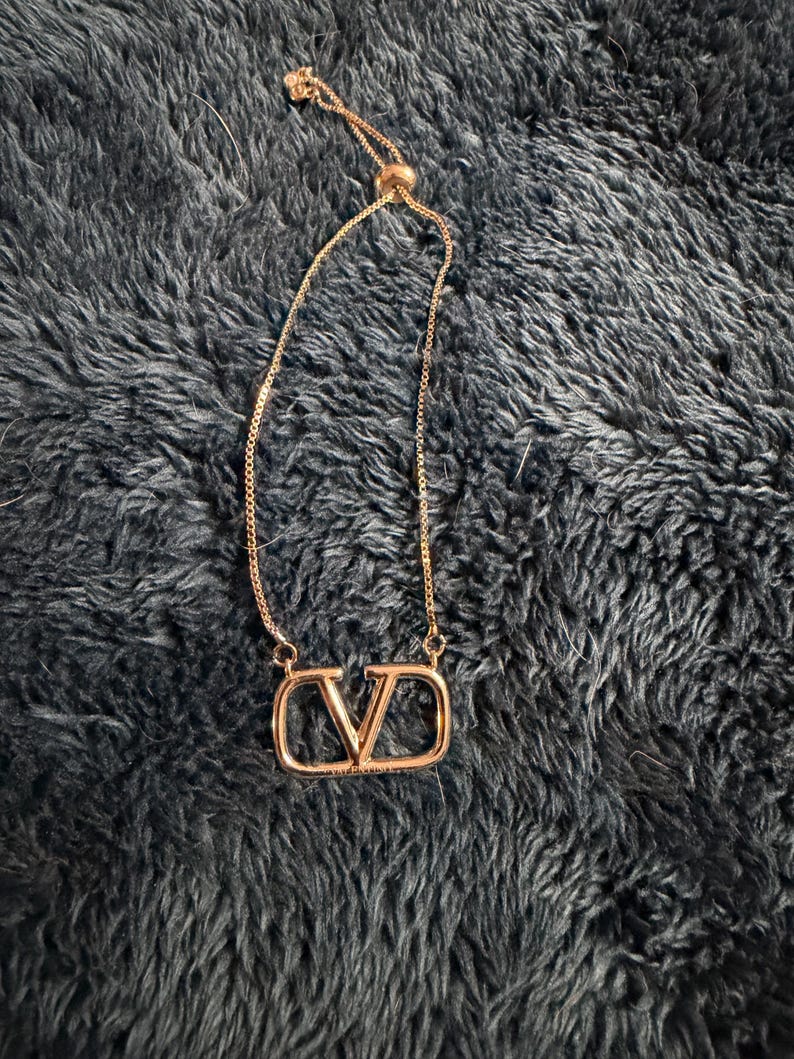 Puede incluir: Un collar de tono oro rosa que presenta un colgante central con forma de letra V dentro de un marco rectangular. La pieza incluye una cadena delicada con un mecanismo de cuenta deslizante ajustable, lo que permite un ajuste personalizado.