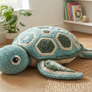 Puede incluir: Un juguete grande de tortuga marina relleno, hecho de hilo de ganchillo en tonos verde azulado, verde oscuro y crema. El caparazón presenta un patrón geométrico de motivos hexagonales, con grandes ojos oscuros y detalles blancos.