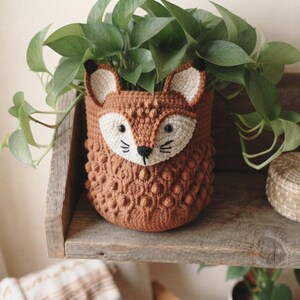 Pode incluir: Uma capa de crochê marrom para vaso de plantas com design de rosto de raposa, detalhes em branco, olhos e nariz bordados em preto. O exterior apresenta um padrão texturizado de pontos pipoca. Uma planta verde está dentro do recipiente cilíndrico.