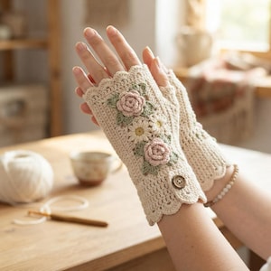 May include: A pair of cream-colored fingerless gloves crafted from crochet yarn. The gloves feature a scalloped edge at the wrist and knuckles, adorned with three-dimensional crocheted pink roses, white daisies, and green leaves, plus a small brown button.
