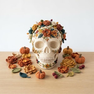Könnte beinhalten: Ein cremefarbener, gehäkelter Totenkopf, verziert mit einem Kranz aus bunten Garnblumen in Orange-, Gelb-, Grün- und Brauntönen. Der Totenkopf ist von einzelnen gehäkelten Blumen, Blättern, Beeren und kleinen Kürbissen umgeben.