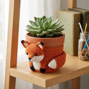 Peut inclure: Un pot de fleurs en terre cuite enveloppé dans une housse au crochet représentant un renard orange et blanc. Le renard a des oreilles à pointes noires, des pattes noires et une queue à pointe blanche. Une plante succulente verte est plantée.