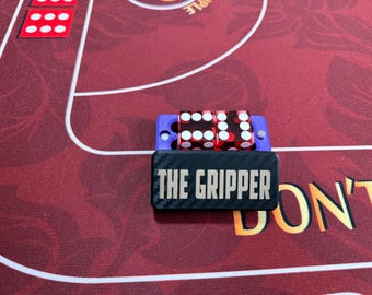 Op de afbeelding: Een zwarte rechthoekige dobbelsteenhouder met een koolstofvezeltextuur en de tekst THE GRIPPER in witte letters. De houder heeft een paarse basis en bevat twee doorschijnende rode dobbelstenen met witte stippen, op een rood speeloppervlak.