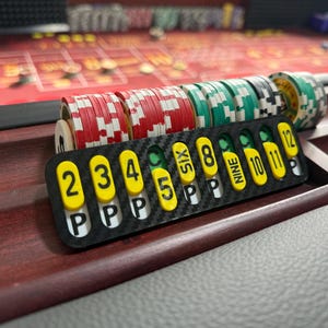May include: A black carbon fiber craps tracker featuring yellow oval markers with black numbers 2, 3, 4, 5, 6, 8, 9, 10, 11, 12 and the letter P printed on them. The tracker is positioned in front of several stacks of red, green, and white casino chips.