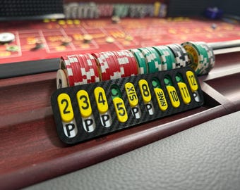Op de afbeelding: Een zwarte koolstofvezel craps-tracker met gele ovale markers met daarop de zwarte cijfers 2, 3, 4, 5, 6, 8, 9, 10, 11, 12 en de letter P. De tracker is geplaatst voor verschillende stapels rode, groene en witte casinofiches.