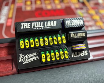 Op de afbeelding: Een zwart craps dobbelsteen trackingbord met koolstofvezeltextuur, voorzien van gele cijfermarkeringen van 2 tot 12. De tekst bevat THE FULL LOAD, THE GRIPPER, THE RAIL RIDERS, The Extreme Gambler en MJGS, met twee kleine paars-gele fiches.