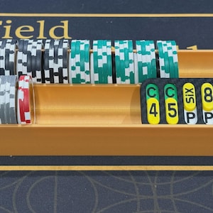 May include: A wooden tray filled with stacks of casino chips in various colors, including black, white, green, and red. A separate black and yellow marker with numbers and the word "PAYS" is also in the tray. The tray is on a black surface with gold text.