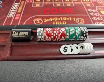 Op de afbeelding: Een houten rek met stapels casinofiches in grijs, rood, groen en zwart, naast een zwarte cilindrische marker met het opschrift THE RAIL RIDERS en verschillende witte ronde fiches met een zwart dollarteken op een craps-tafel.