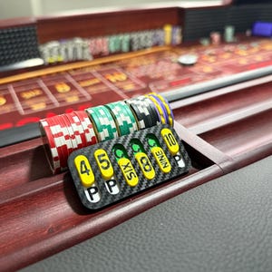 May include: A craps table marker featuring a carbon fiber pattern with yellow and white rectangular inserts labeled 4, 5, SIX, 8, NINE, and 10, positioned next to a stack of red, green, black, and purple casino chips on a wooden surface.