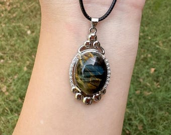 May include: A pendant necklace featuring an oval-shaped tiger's eye gemstone with golden and dark blue bands. The stone is set in a silver-toned metal frame adorned with small clear crystals, suspended from a simple black cord necklace.