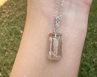 May include: A silver-toned chain necklace featuring a rectangular, translucent, light brown resin pendant. The pendant is suspended from a small silver-toned bail adorned with three small, clear, round-cut stones that connect the chain to the pendant.