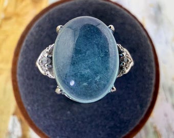 May include: A silver ring featuring a large, oval-shaped, translucent blue cabochon gemstone set in the center. The metal band is decorated with small, clear accent stones on both sides of the central setting, creating a detailed, symmetrical design.