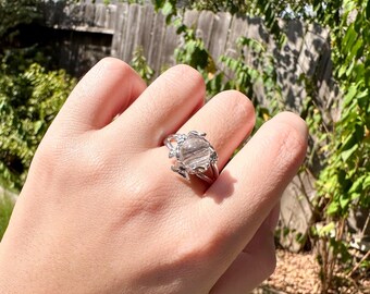 May include: A silver-toned ring featuring a round, translucent gemstone with visible dark, needle-like inclusions. The setting has a decorative, organic metal design that wraps around the base of the stone, securing it to the polished band.