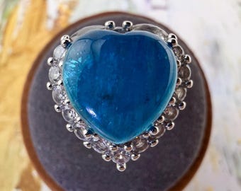 May include: A ring featuring a large, heart-shaped, translucent blue gemstone set in a silver-toned metal band. The central blue stone is surrounded by a halo of small, clear, round-cut white stones that follow the heart-shaped outline of the setting.