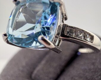 May include: A silver-toned ring featuring a large, cushion-cut light blue gemstone held by four prongs. The band is accented with three small, square-cut clear stones embedded along each side of the shank, creating a bright, reflective appearance.