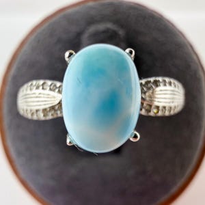 May include: A silver ring featuring a large, oval-shaped, light blue Larimar gemstone held by four prongs. The band of the ring is crafted from silver and displays a textured, leaf-like pattern along the sides, adding detail to the metalwork.
