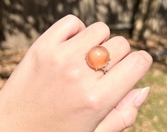May include: A ring featuring a large, smooth, round, peach-colored gemstone set in a metallic band. The stone has a translucent, glowing appearance with a subtle shimmer, and the band is detailed with small, decorative metal accents around the base.