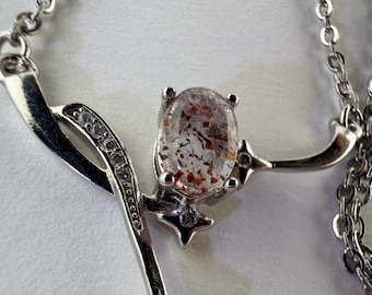 May include: A silver-toned necklace featuring a pendant with an oval-shaped, clear stone containing reddish-brown inclusions. The pendant has a curved, metallic frame accented with small, clear, round stones and a star-shaped detail at the bottom.