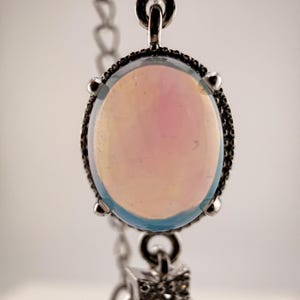 May include: A silver-toned bracelet featuring a central oval cabochon stone with a pale iridescent pink and blue glow, held by four prongs. Below the main stone, a small star-shaped charm encrusted with tiny clear stones hangs from a silver chain.