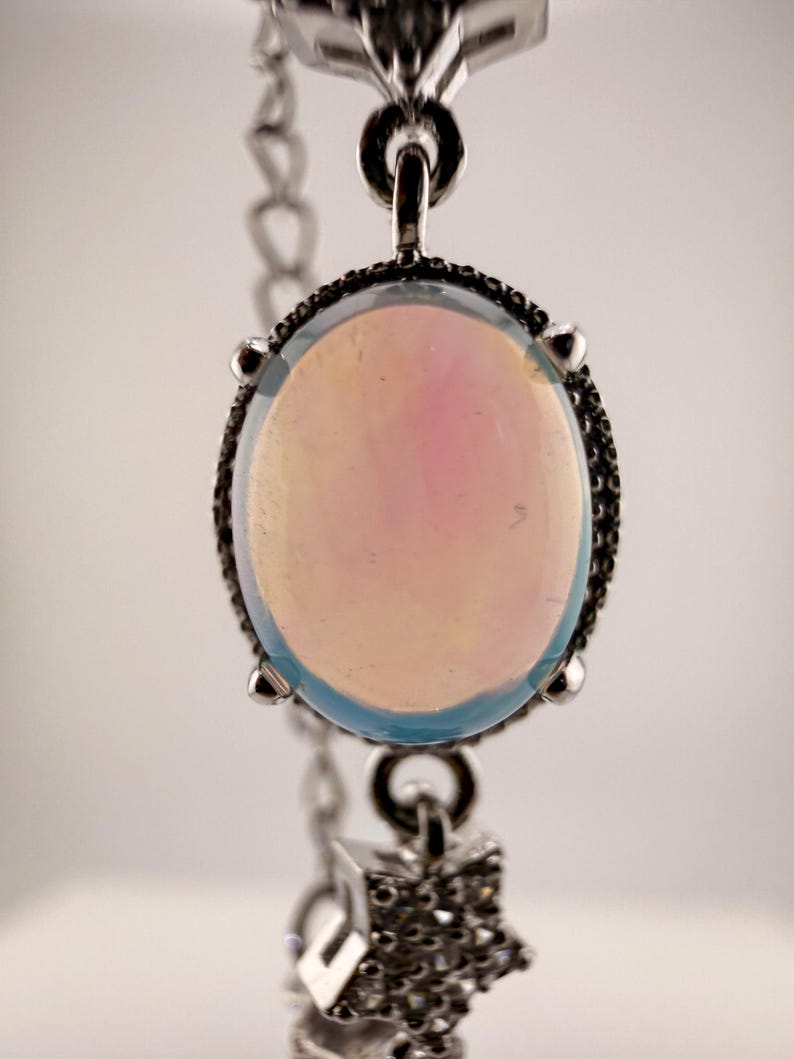 May include: A silver-toned bracelet featuring a central oval cabochon stone with a pale iridescent pink and blue glow, held by four prongs. Below the main stone, a small star-shaped charm encrusted with tiny clear stones hangs from a silver chain.