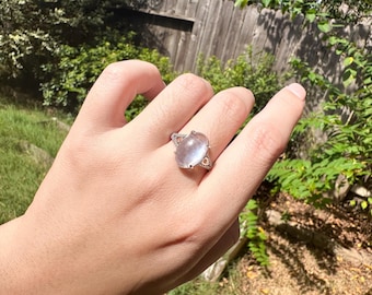 May include: A silver-toned ring featuring a large, oval-shaped, translucent pale pink cabochon stone set in a decorative metal band with small, triangular cut-out details on the sides and a textured, beaded edge along the shank of the ring.