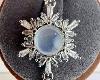 May include: A silver-toned pendant shaped like a snowflake, featuring a central round, translucent, pale blue cabochon stone surrounded by clear, baguette-cut and round-cut crystals. The pendant includes a decorative bail and a small dangling charm.