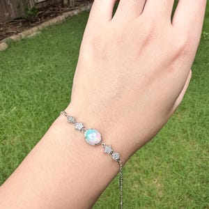 May include: A silver-toned chain bracelet featuring a central oval-shaped iridescent opal-like stone surrounded by small clear stones. The bracelet includes two star-shaped accents and two circular accents, each set with small clear stones on either side.