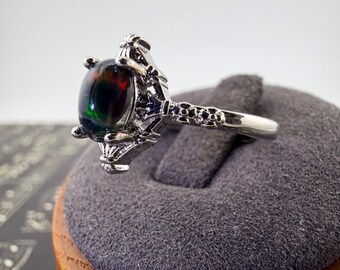 May include: A silver-toned ring featuring a dark, oval-shaped cabochon stone with iridescent flashes of green and red. The metal band is detailed with a textured, claw-like setting that secures the central stone, showcasing a polished metallic finish.