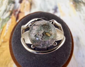 May include: A silver ring featuring a circular, translucent cabochon stone with internal inclusions in shades of green, grey, and purple. The metal band has a textured, organic design that curves around the central stone, securing it in place.