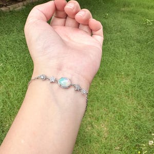 May include: A silver-toned chain bracelet featuring a central oval-shaped iridescent opal stone set in a decorative frame, flanked by small flower-shaped accents and clear round stones, secured with a delicate link chain around the wrist.