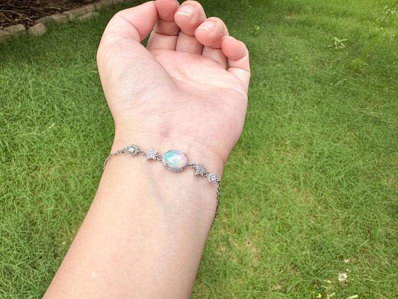 May include: A silver-toned chain bracelet featuring a central oval-shaped iridescent opal stone set in a decorative frame, flanked by small flower-shaped accents and clear round stones, secured with a delicate link chain around the wrist.