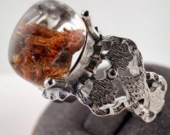 May include: A silver-toned ring featuring a large, smooth, clear cabochon stone with internal reddish-brown, fibrous inclusions. The band is crafted with an intricate, textured, openwork metal design that wraps around the finger.