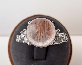 May include: A silver-toned ring featuring a large, round, cabochon-cut rutilated quartz gemstone with visible reddish-brown needle-like inclusions. The band is accented with small, clear, sparkling stones on both sides of the central setting.