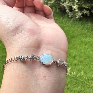 May include: A silver-toned bracelet featuring a central oval-shaped iridescent opal stone surrounded by small clear stones. The chain includes star-shaped charms encrusted with tiny clear stones and a delicate link design with a secure clasp mechanism.
