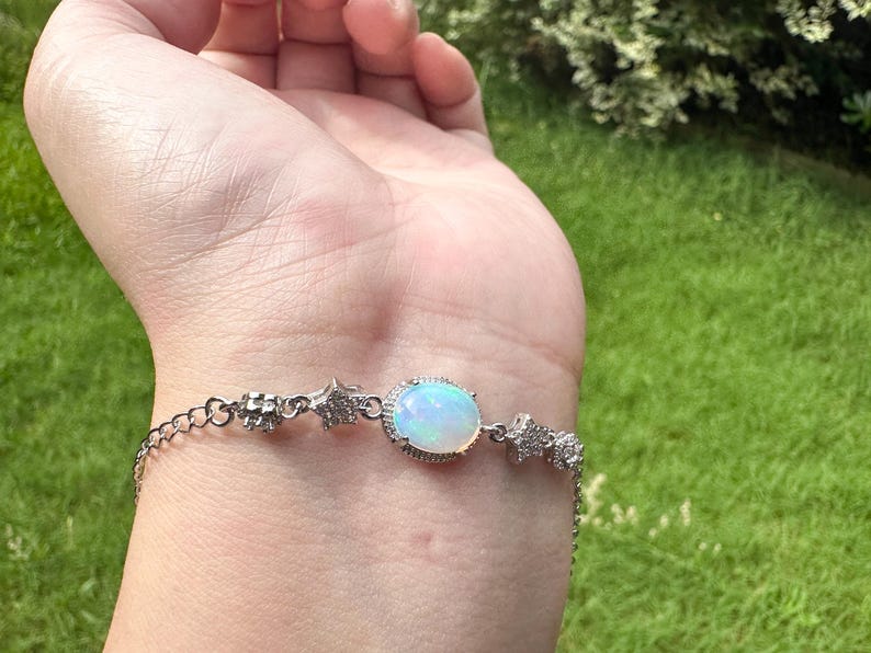 May include: A silver-toned bracelet featuring a central oval-shaped iridescent opal stone surrounded by small clear stones. The chain includes star-shaped charms encrusted with tiny clear stones and a delicate link design with a secure clasp mechanism.