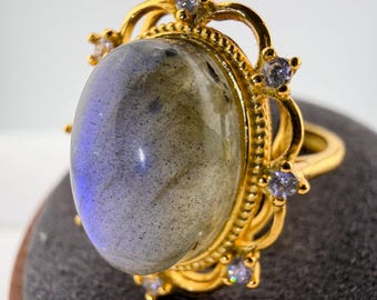 May include: A gold ring featuring a large, oval-shaped labradorite cabochon with blue iridescence. The setting is decorated with a scalloped metal border and small, clear, round-cut accent stones placed at intervals around the central gemstone.